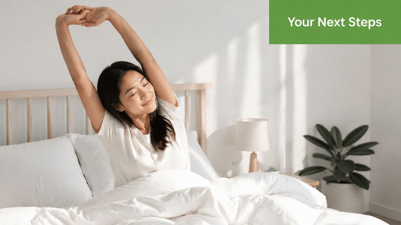 A young Asian woman smiling and stretching her arms while waking up in a bright, cozy bedroom.